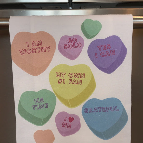 A white tea towel with candy hearts and self-love phrases hangs in a kitchen