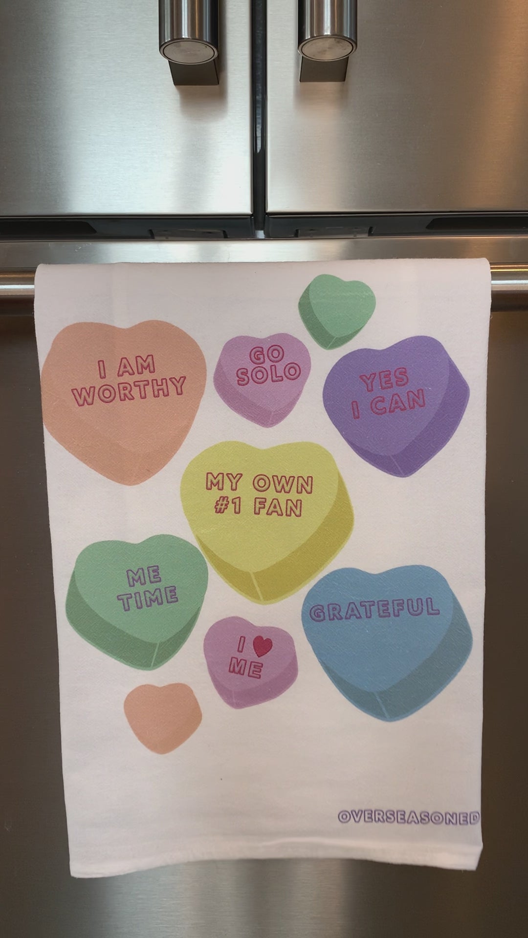 A white tea towel with candy hearts and self-love phrases hangs in a kitchen