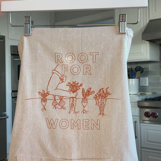 A natural chambray apron with orange block letters that read "Root for Women" hangs on a hanger