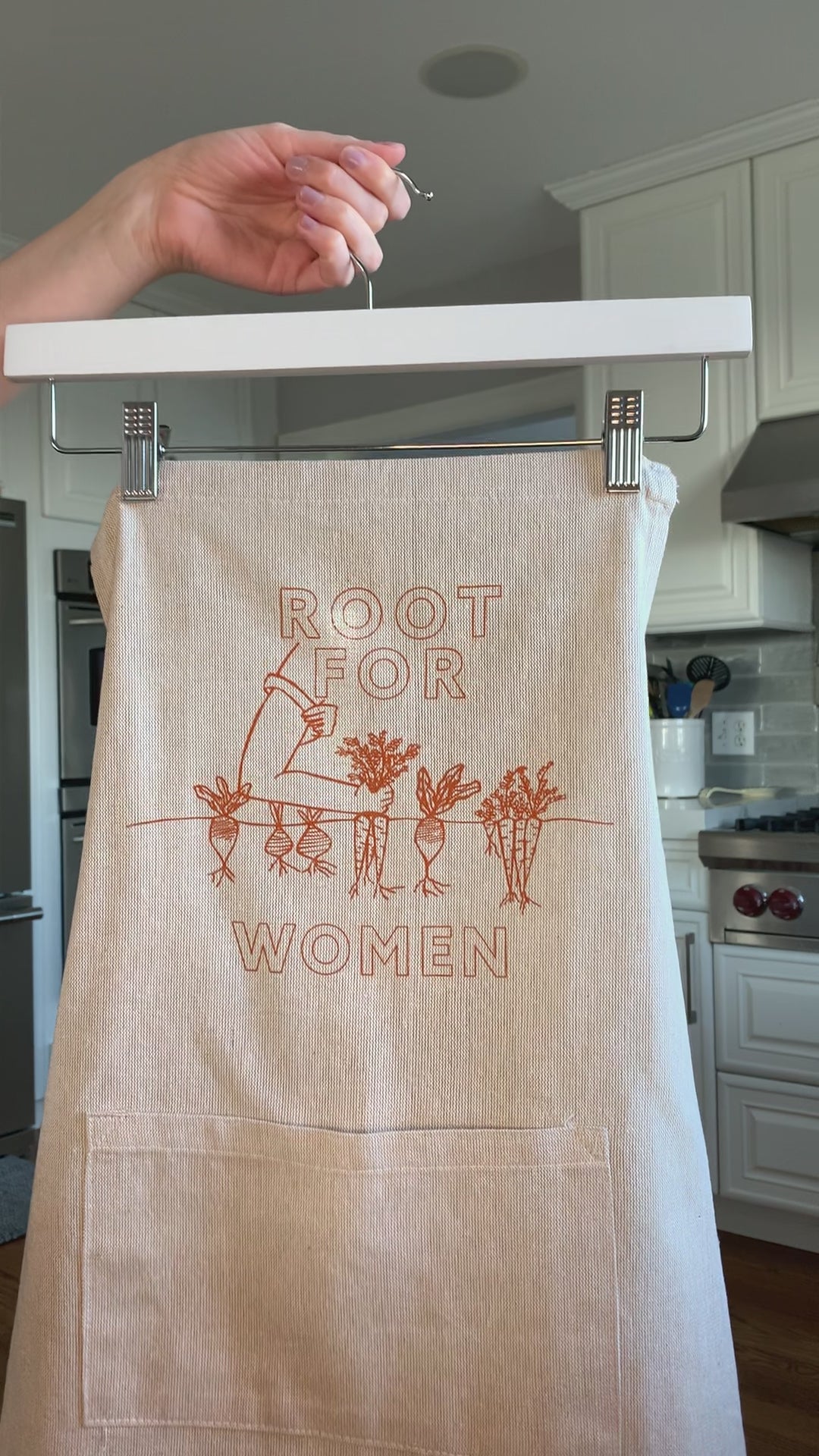 A natural chambray apron with orange block letters that read "Root for Women" hangs on a hanger