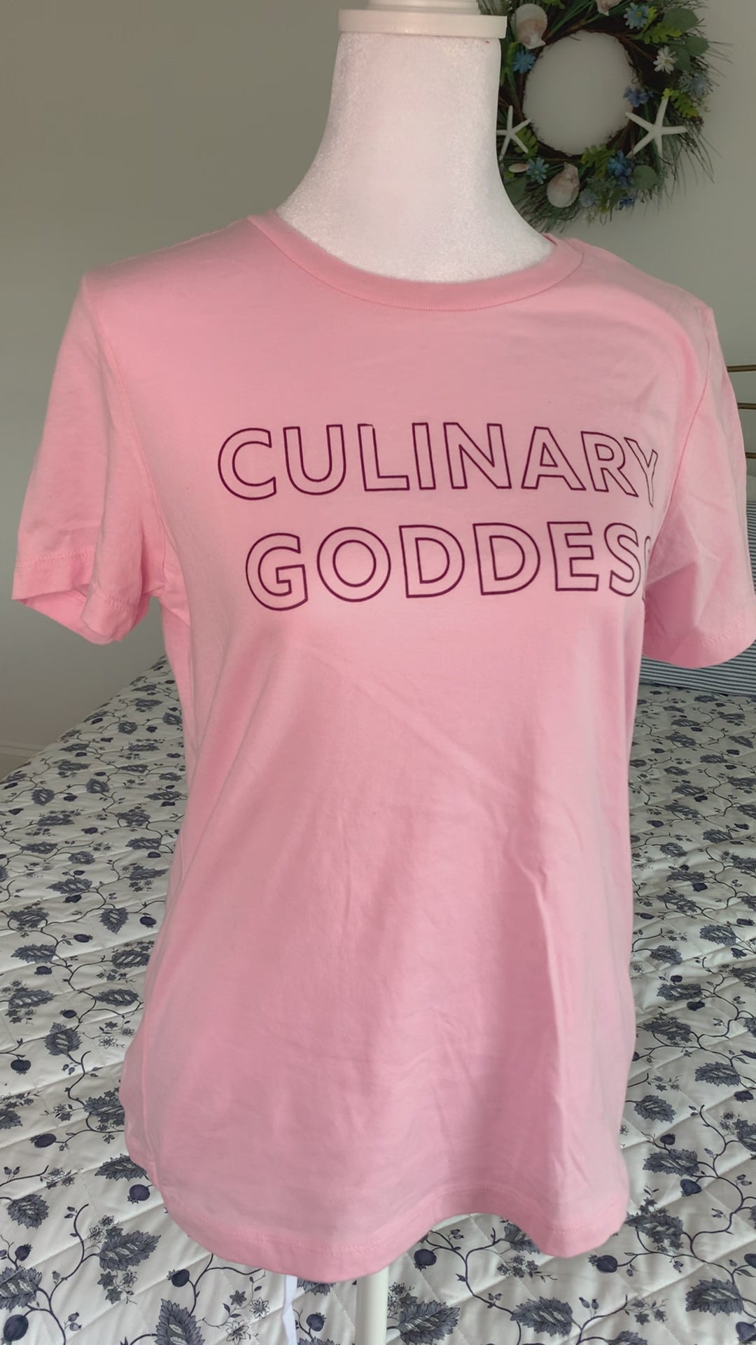 A light pink women's t-shirt that reads "Culinary Goddess" hangs on a manikin