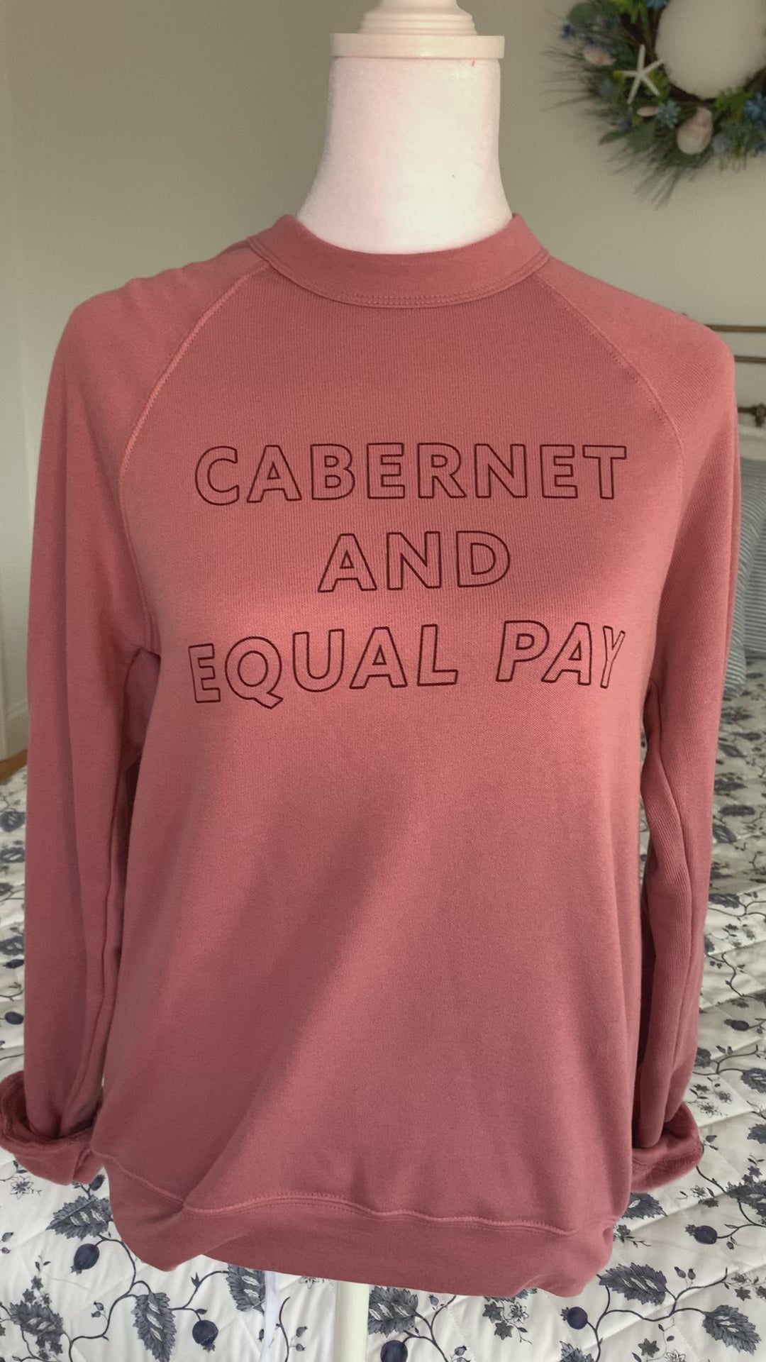 A mauve crewneck that reads "Cabernet and Equal Pay" hangs on a manikin 