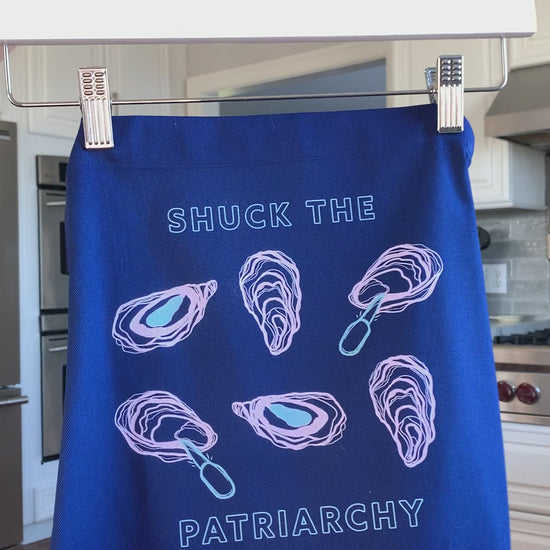 A blue apron that reads "Shuck the Patriarchy" with oyster illustrations hangs on a hanger