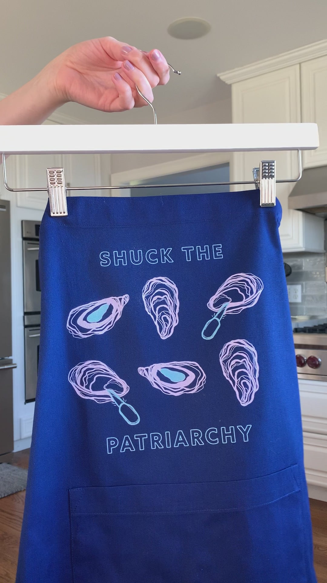 A blue apron that reads "Shuck the Patriarchy" with oyster illustrations hangs on a hanger