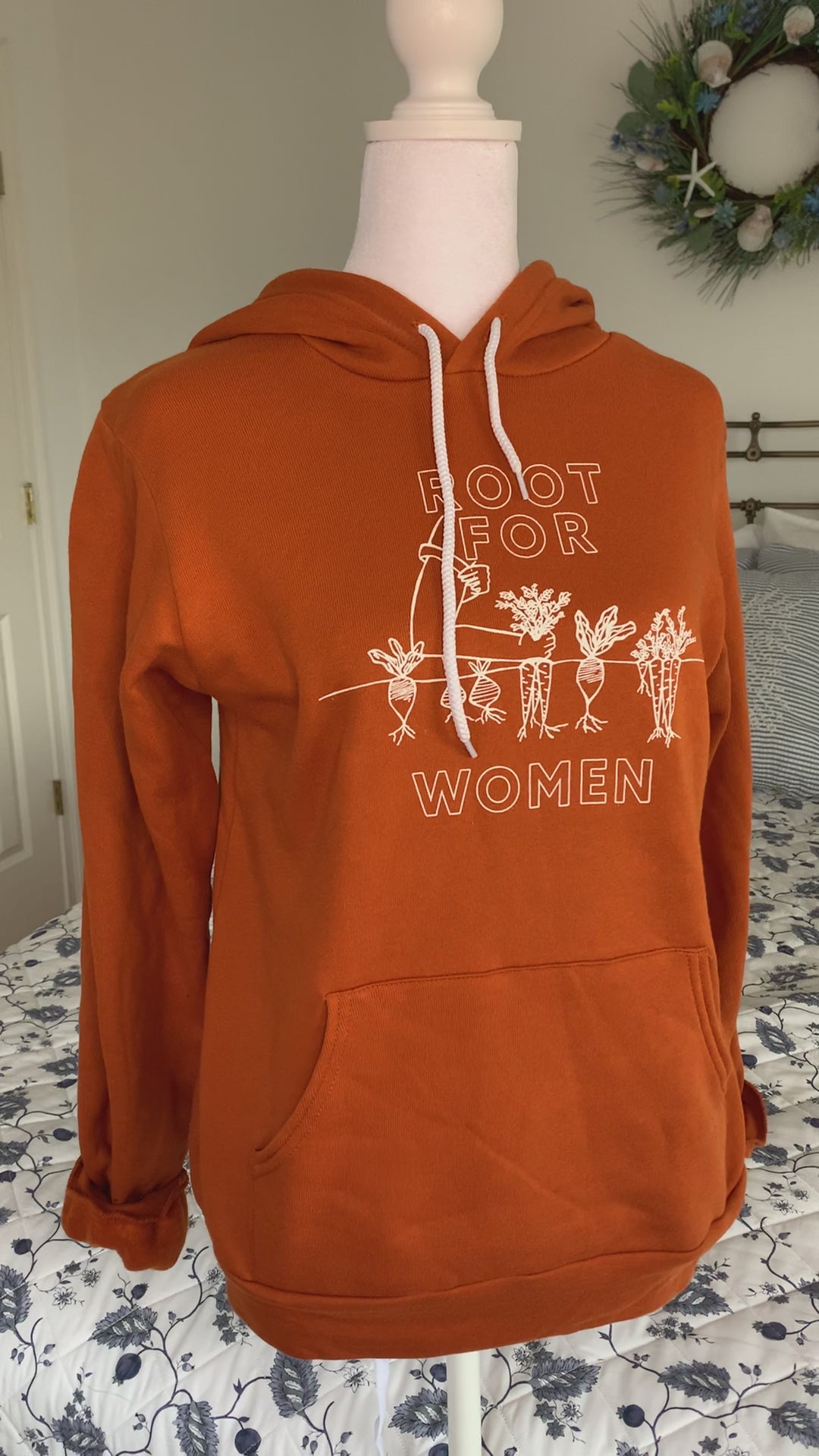 An orange hoodie that reads "Root for Women" hangs on a manikin