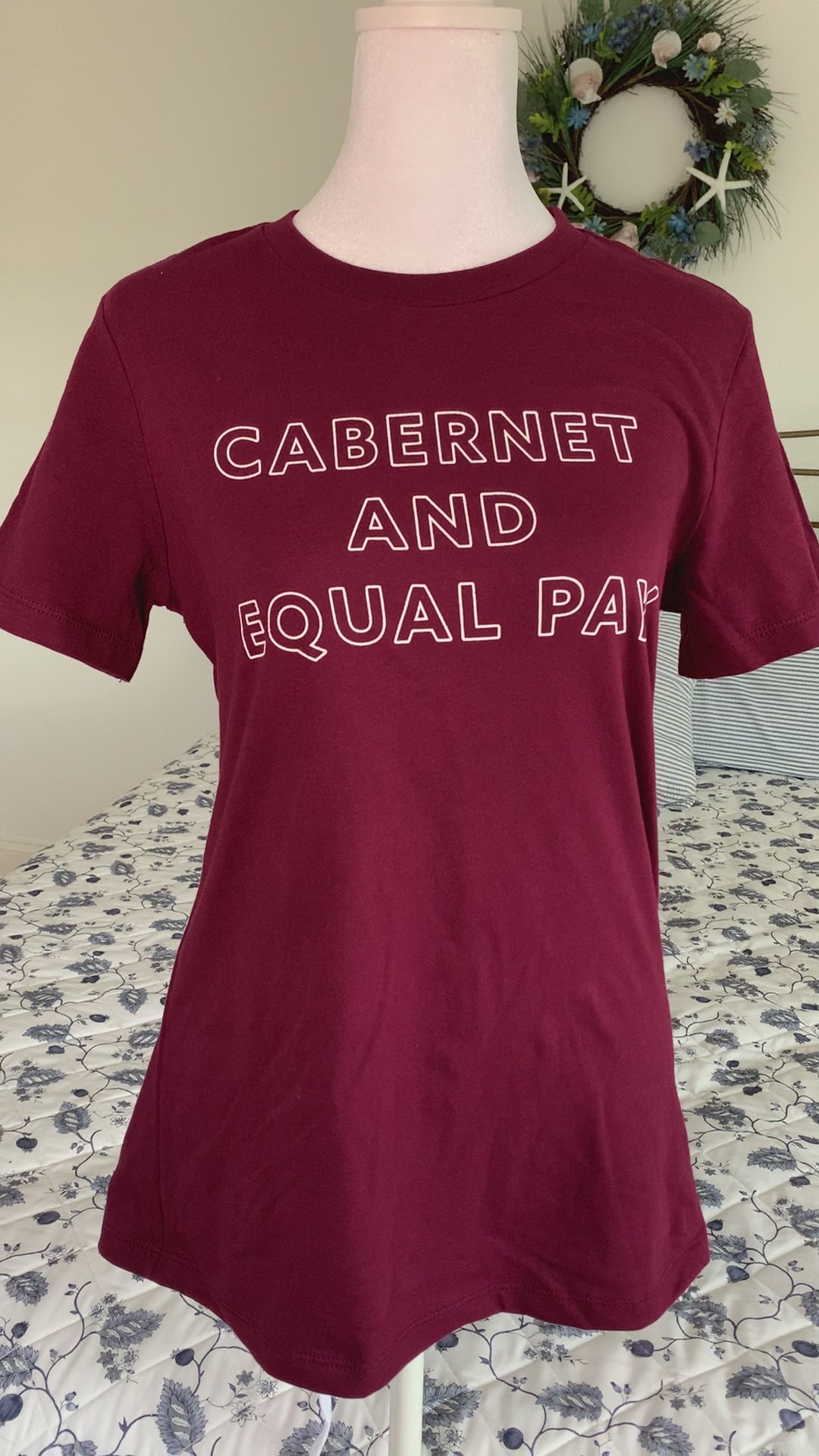 A maroon t-shirt that reads "Cabernet and Equal Pay" hangs on a manikin
