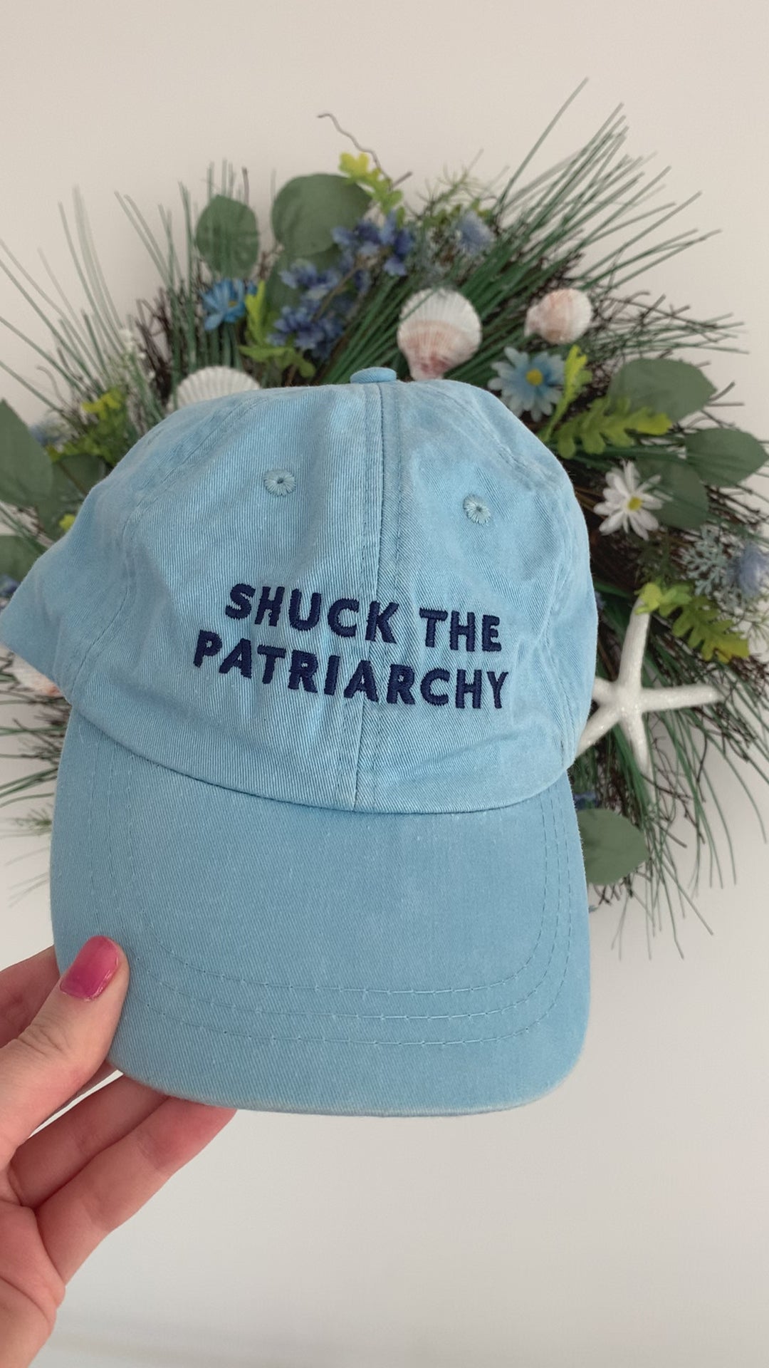 A woman holds a light blue baseball hat that reads "Shuck the Patriarchy"
