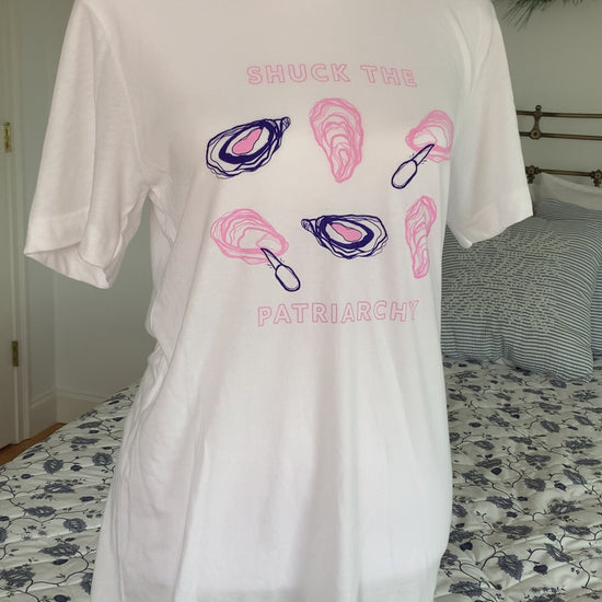 A white t-shirt that reads "Shuck the Patriarchy" in pink block letters with oyster illustrations hangs on a manikin