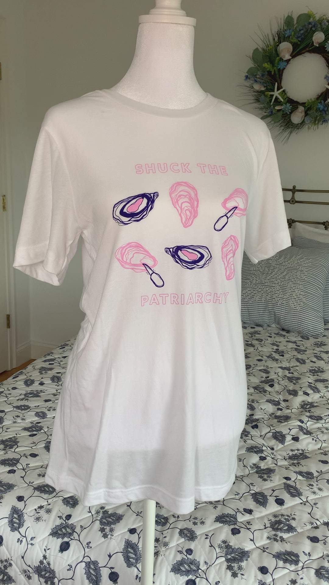 A white t-shirt that reads "Shuck the Patriarchy" in pink block letters with oyster illustrations hangs on a manikin