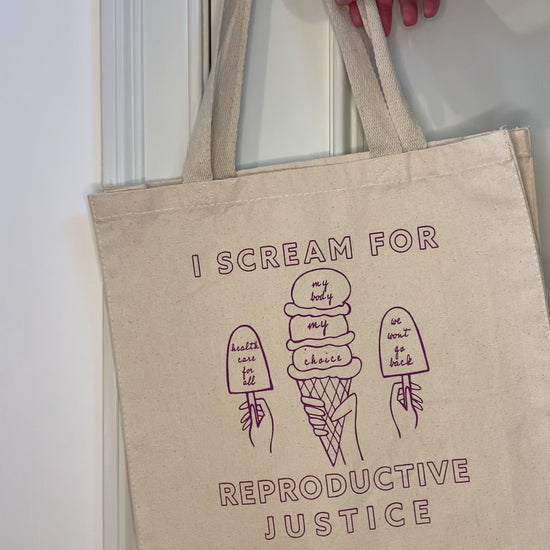 A canvas tote with raspberry block letters that reads "I scream for reproductive justice" hangs on a doorknob