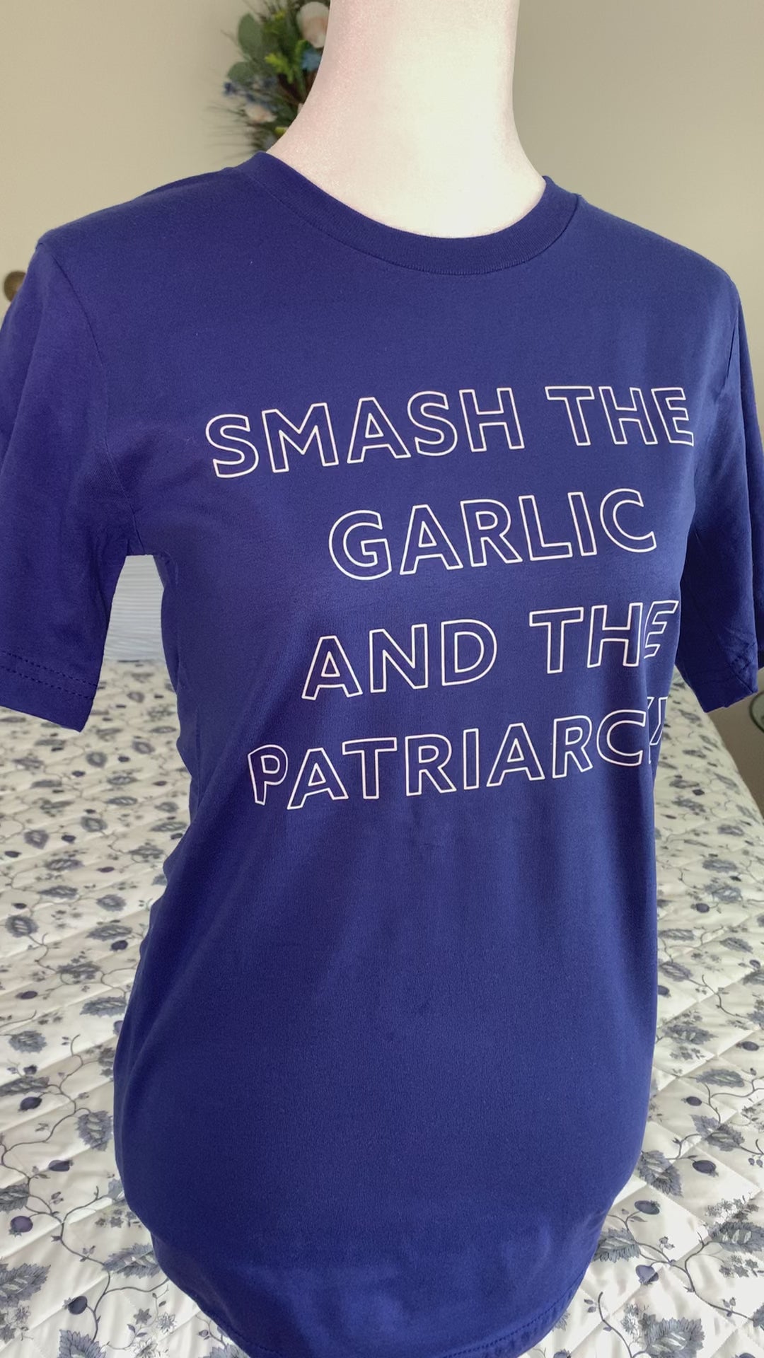 A bright blue tee that reads "Smash the Garlic and the Patriarchy" in white block letters hangs on a manikin