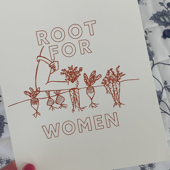 A woman holds print that reads "Root for Women" in orange block letters with a garden illustration 