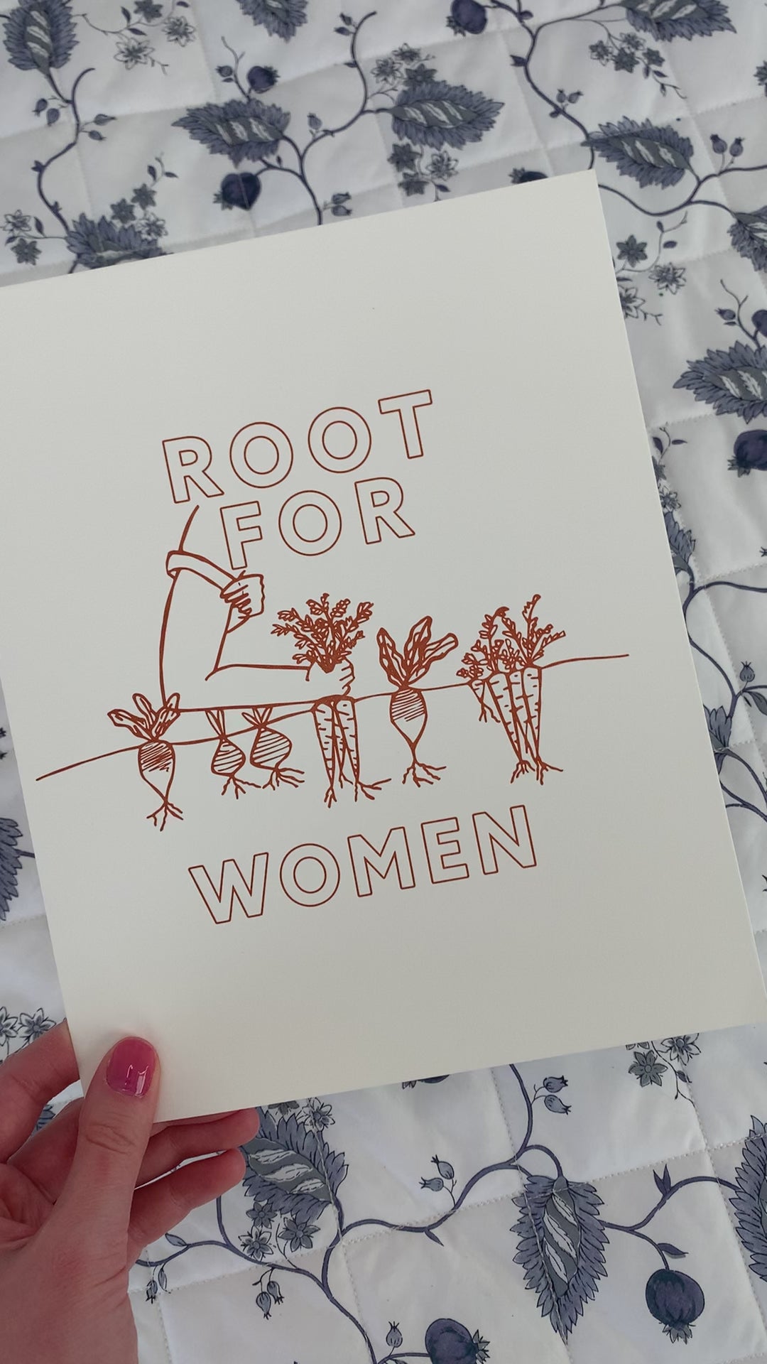 A woman holds print that reads "Root for Women" in orange block letters with a garden illustration 