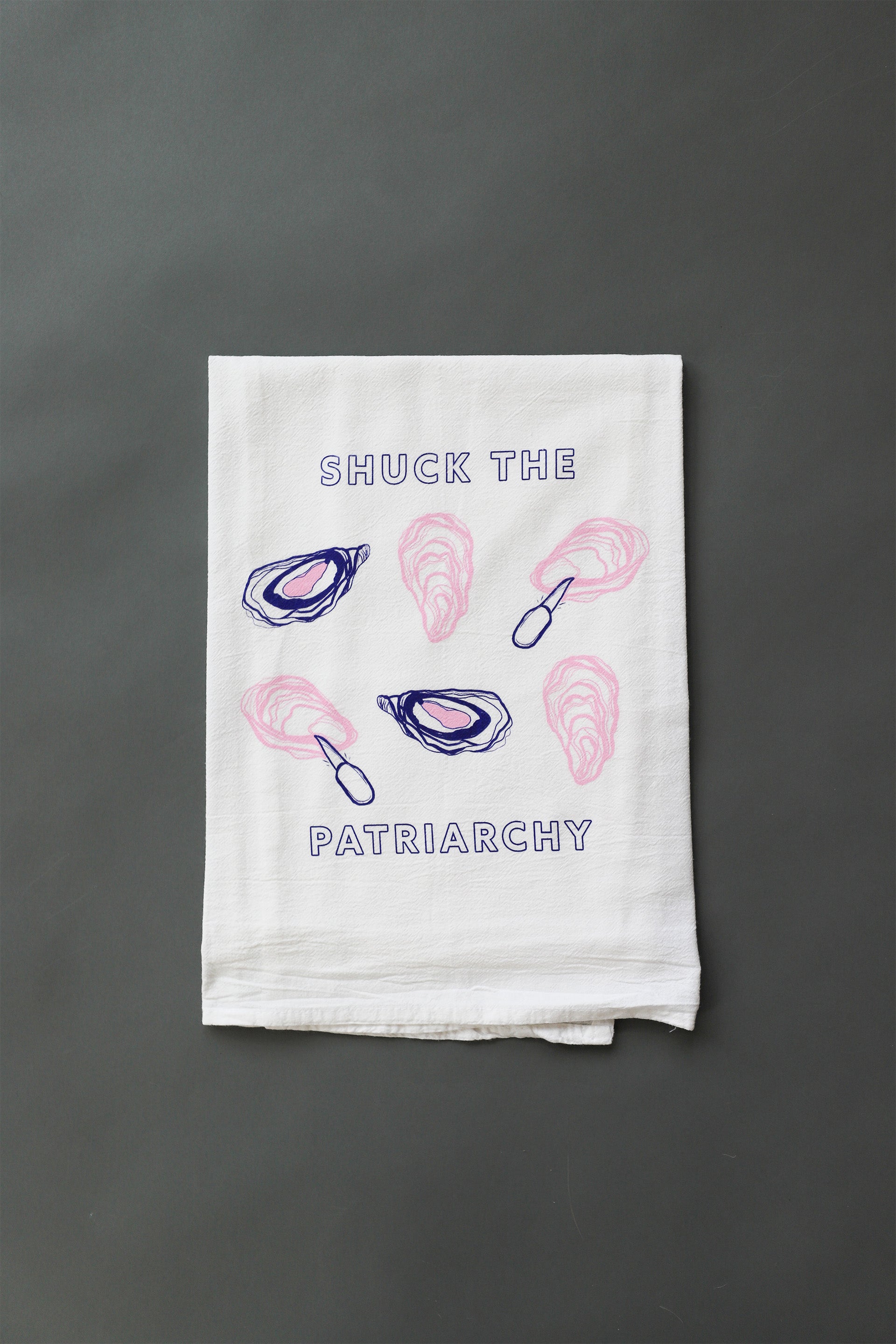 A white tea towel with "Shuck the Patriarchy" in blue block letters and pink and blue oyster illustrations 