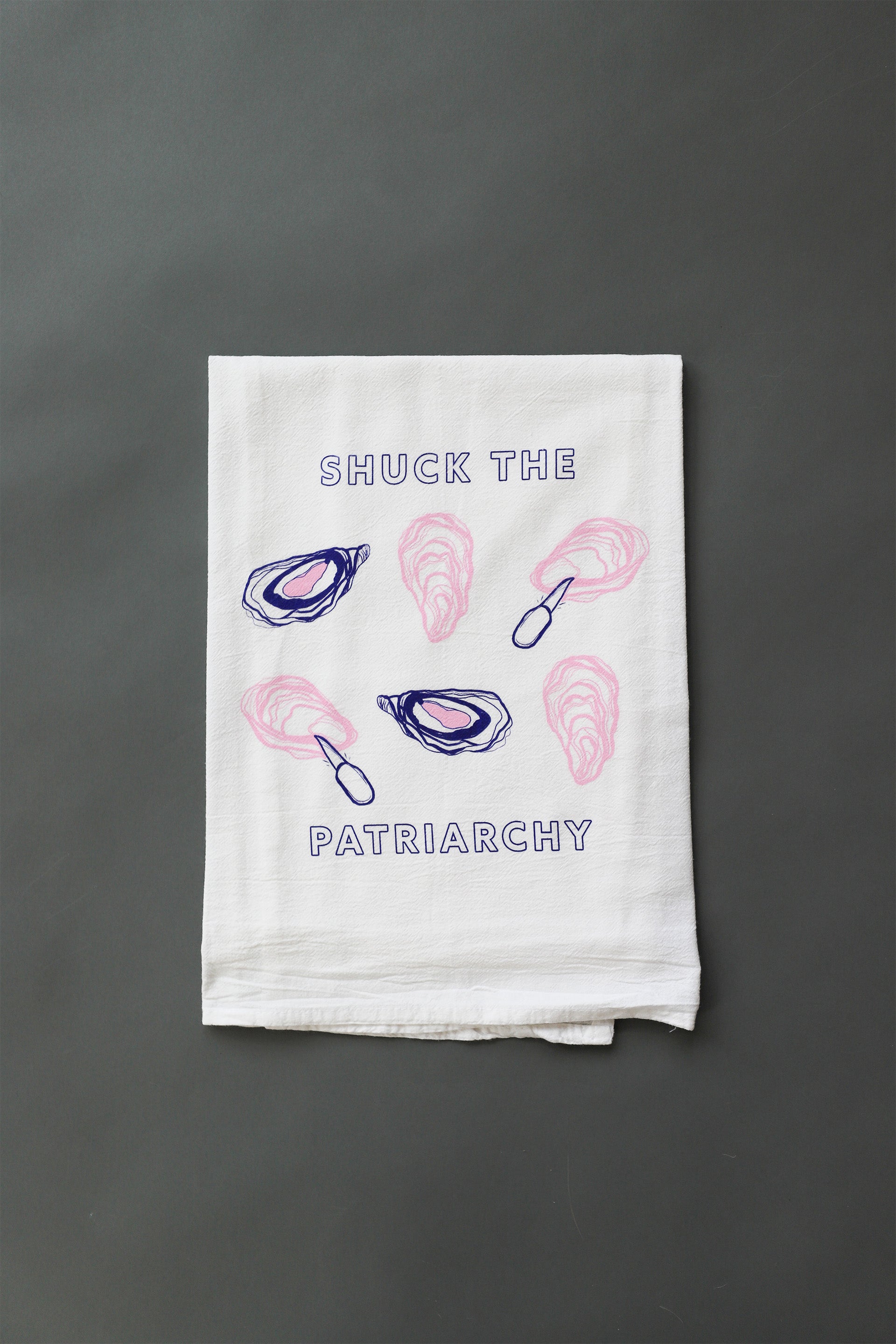 A white tea towel with "Shuck the Patriarchy" in blue block letters and pink and blue oyster illustrations 