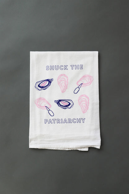 A white tea towel with "Shuck the Patriarchy" in blue block letters and pink and blue oyster illustrations 
