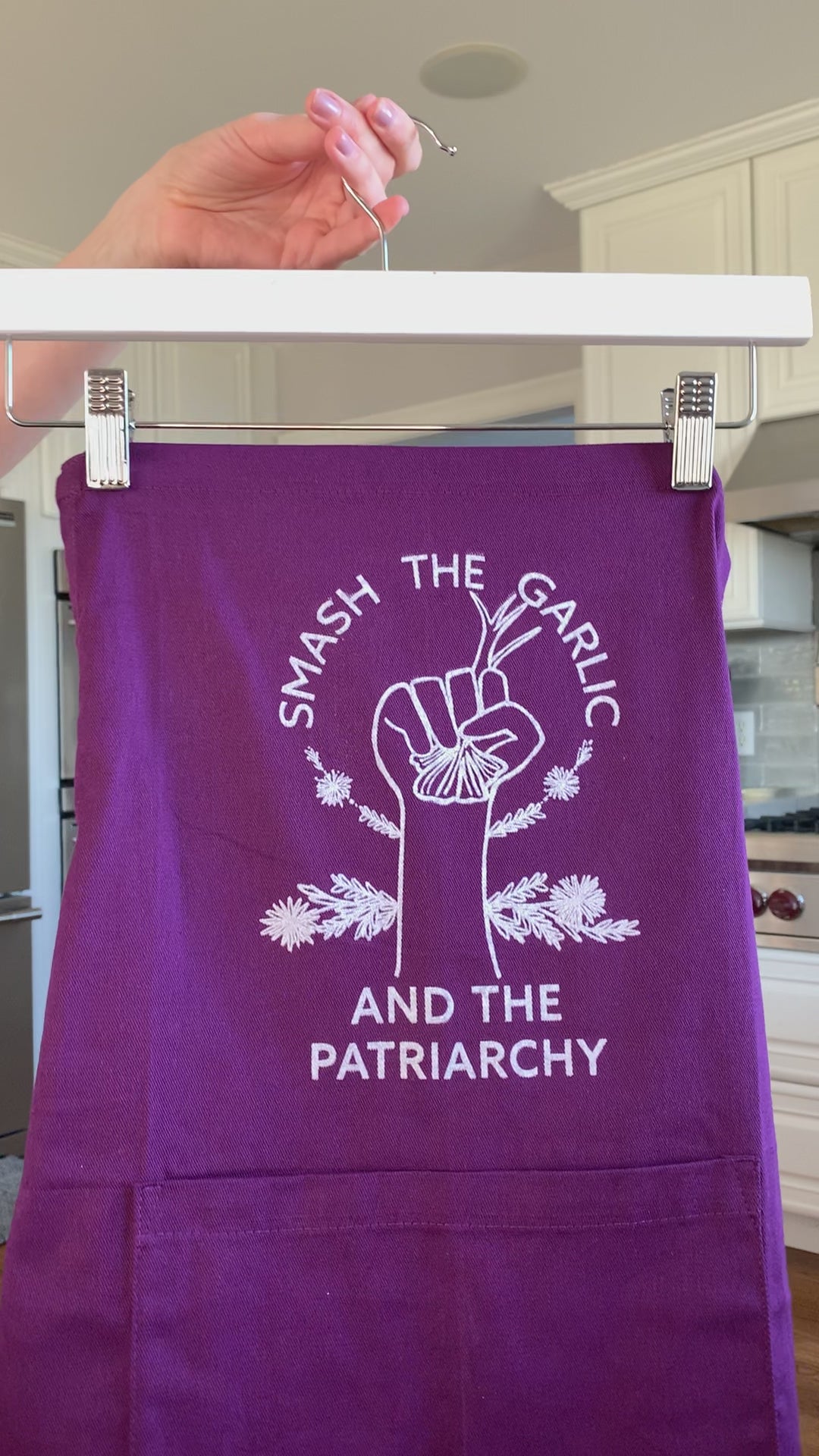 A dark purple "Smash the Garlic and the Patriarchy" plus size apron hangs on a hanger