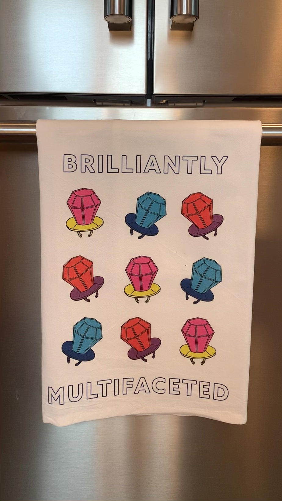 A white tea towel that reads "Brilliantly Multifaceted" hangs in a kitchen