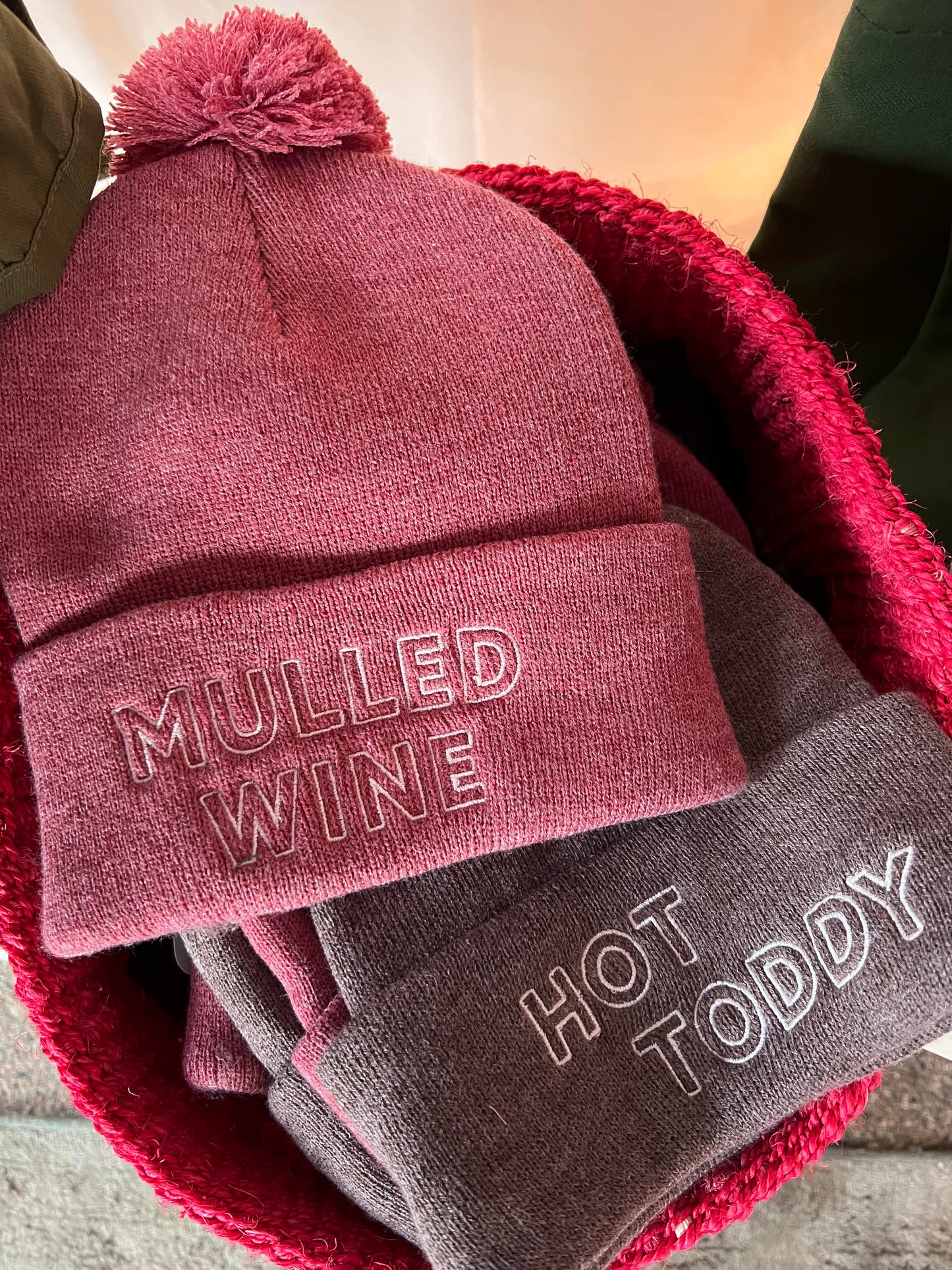 A pink beanie with the words Mulled Wine in white embroidery and a brown beanie with the words Hot Toddy in white embroidery