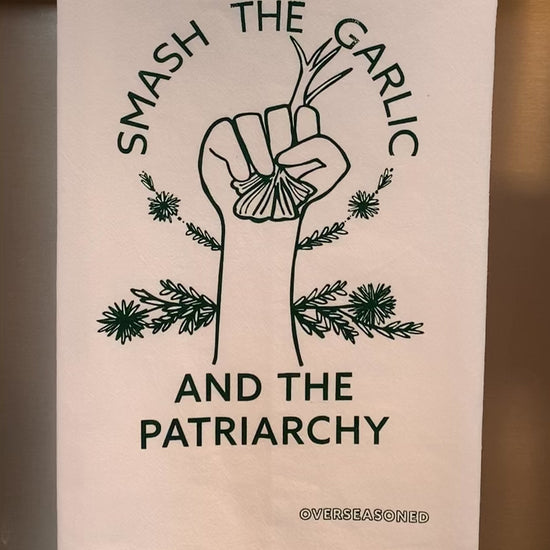 A white tea towel with green lettering that reads "Smash the Garlic and the Patriarchy" and a garlic illustration hangs in a kitchen