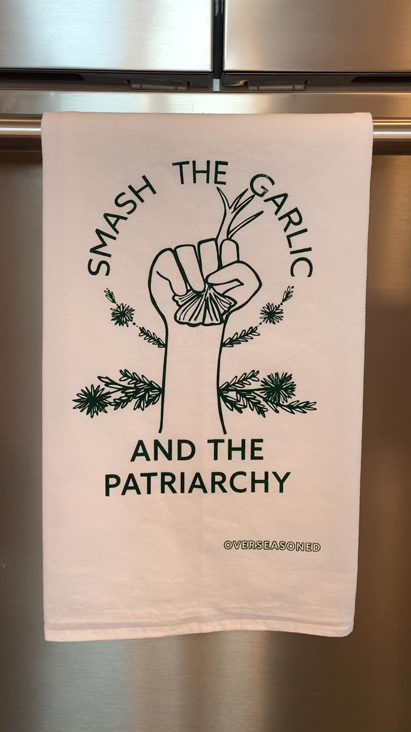 A white tea towel with green lettering that reads "Smash the Garlic and the Patriarchy" and a garlic illustration hangs in a kitchen