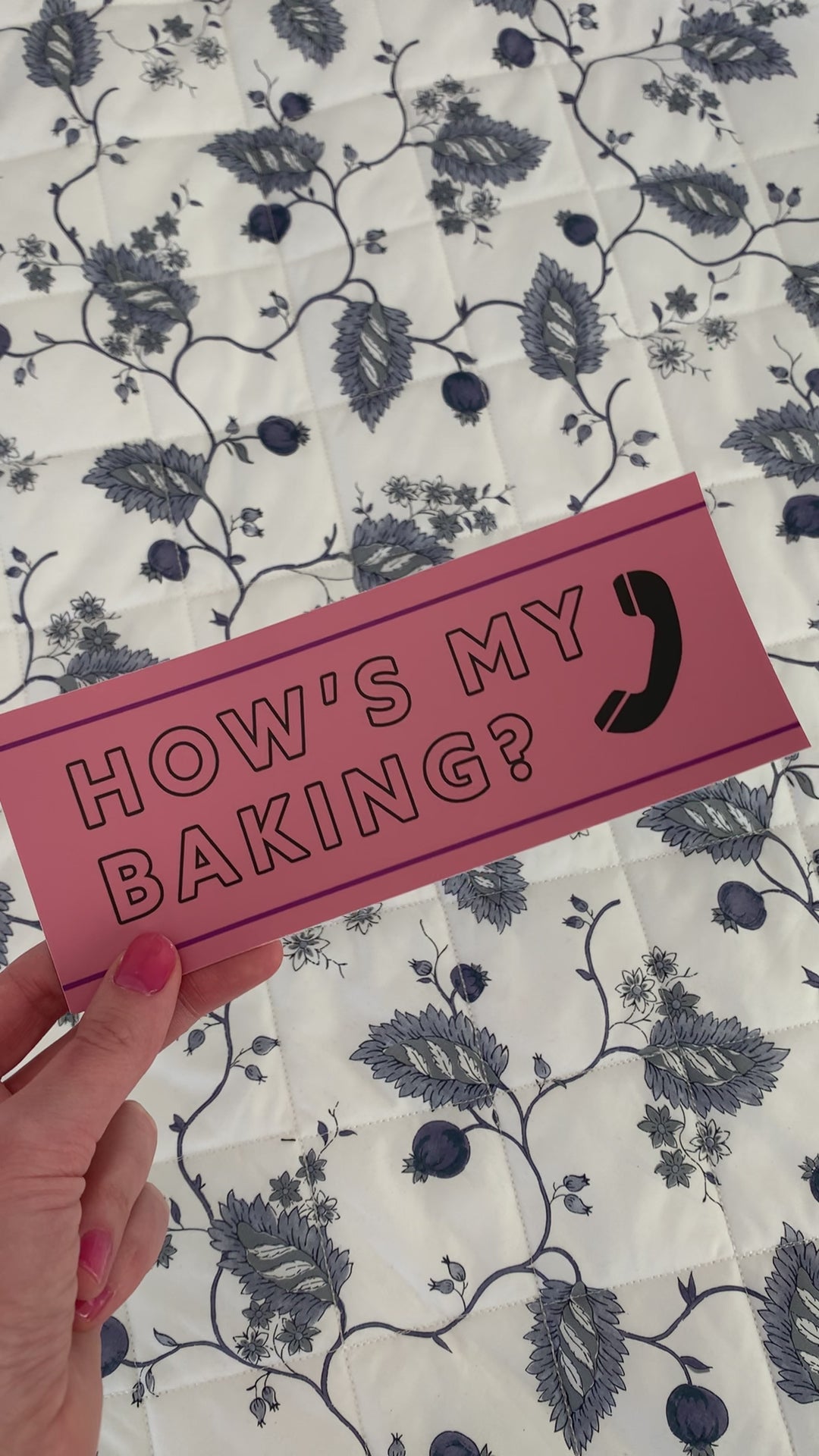 A woman holds a bumper sticker that reads "Hows my baking?"