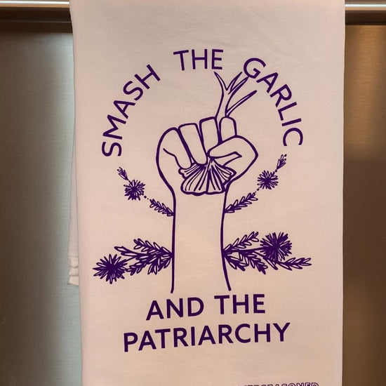 A white tea towel with purple lettering that reads "Smash the Garlic and the Patriarchy" and a garlic design hangs in a kitchen