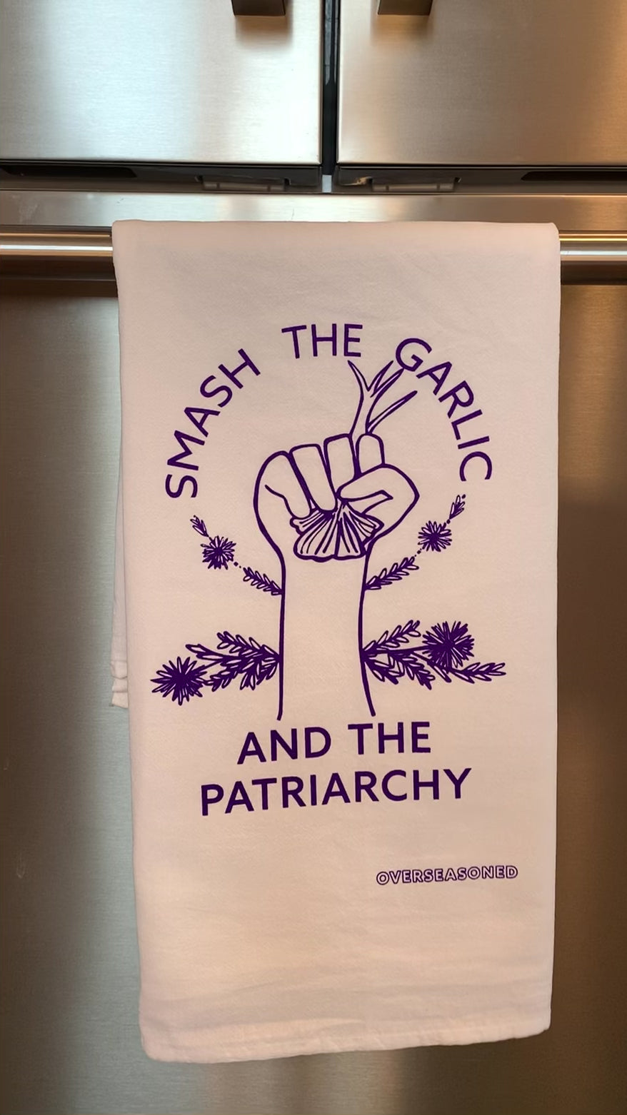 A white tea towel with purple lettering that reads "Smash the Garlic and the Patriarchy" and a garlic design hangs in a kitchen