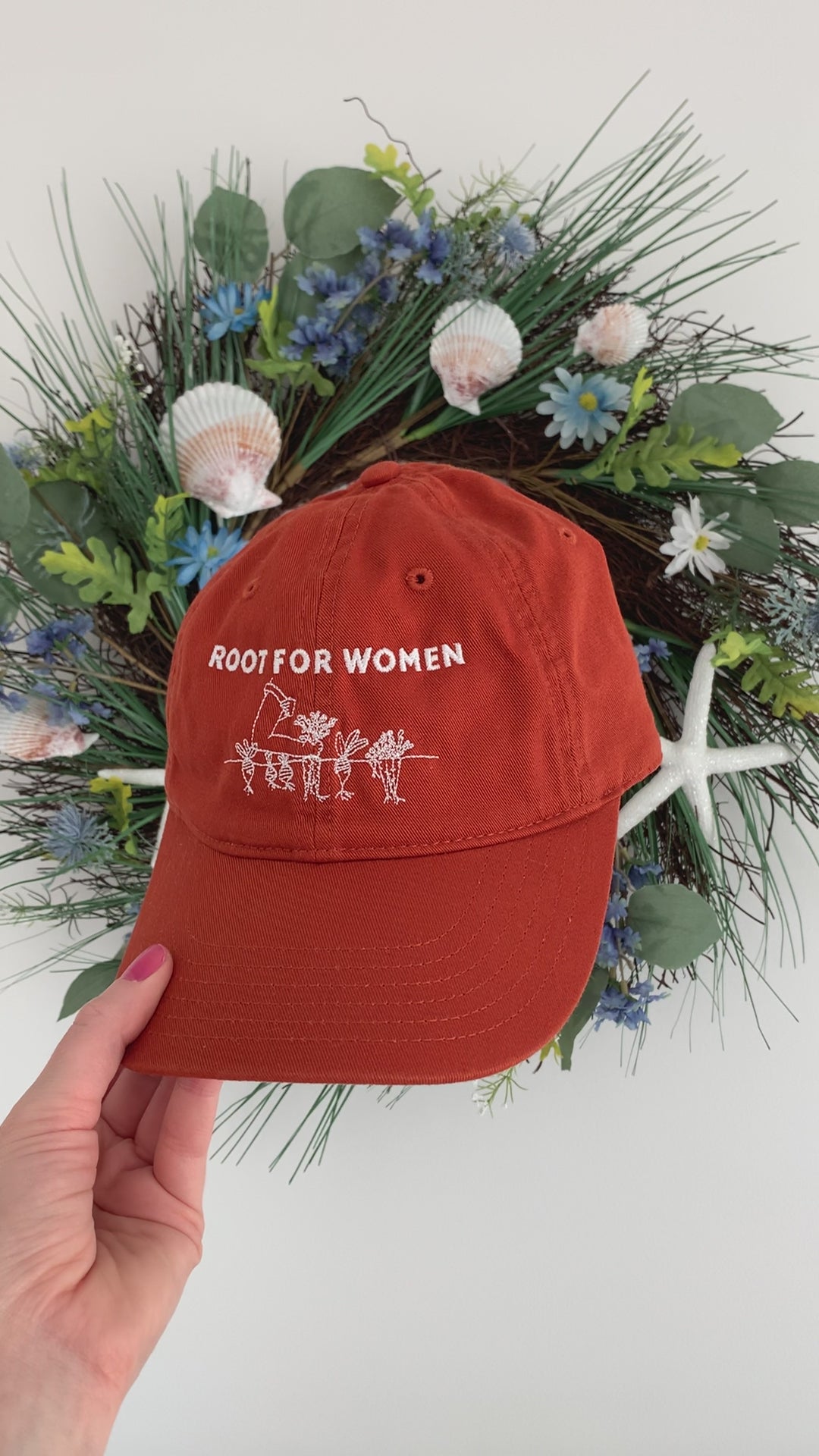 A woman holds an orange baseball hat with with words "Root for Women" embroidered in white