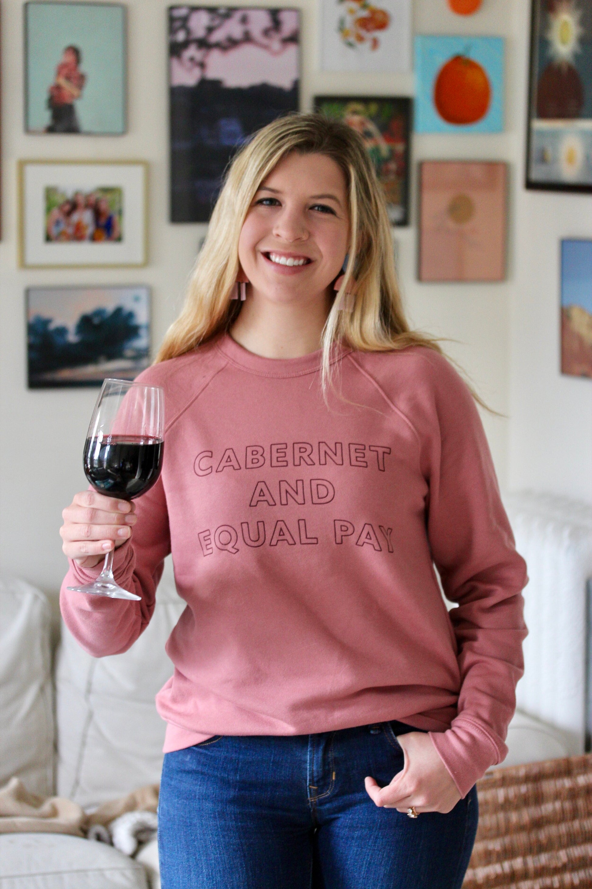 Woman wearing pink sweatshirt with the words "Cabernet and Equal Pay" holding glass of wine