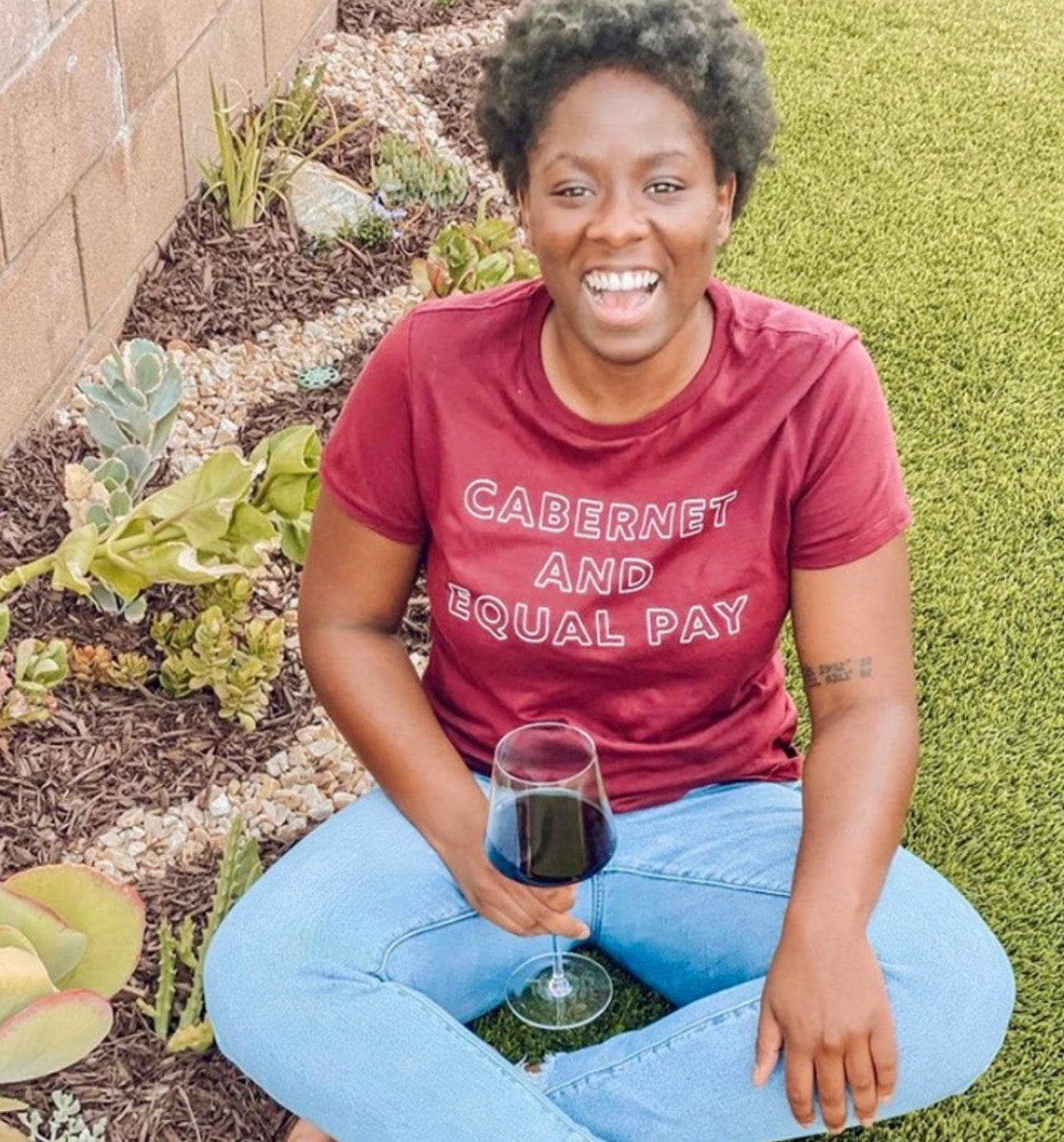 A woman sitting in the grass with a red shirt that says "Cabernet and Equal Pay" and a glass of wine