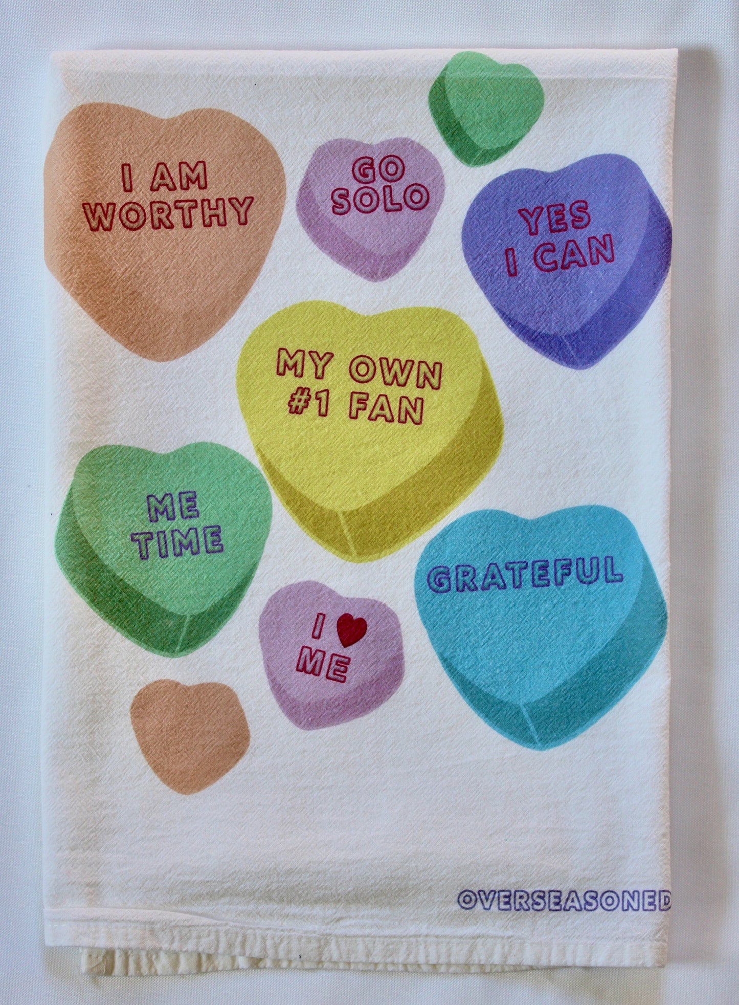 White tea towel with multicolored candy heart design with self-love phrases