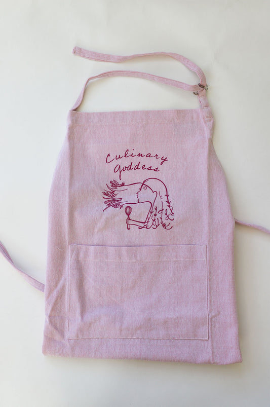 Pink chambray apron with the words "Culinary Goddess" in cursive and illustration of a woman with a whisk