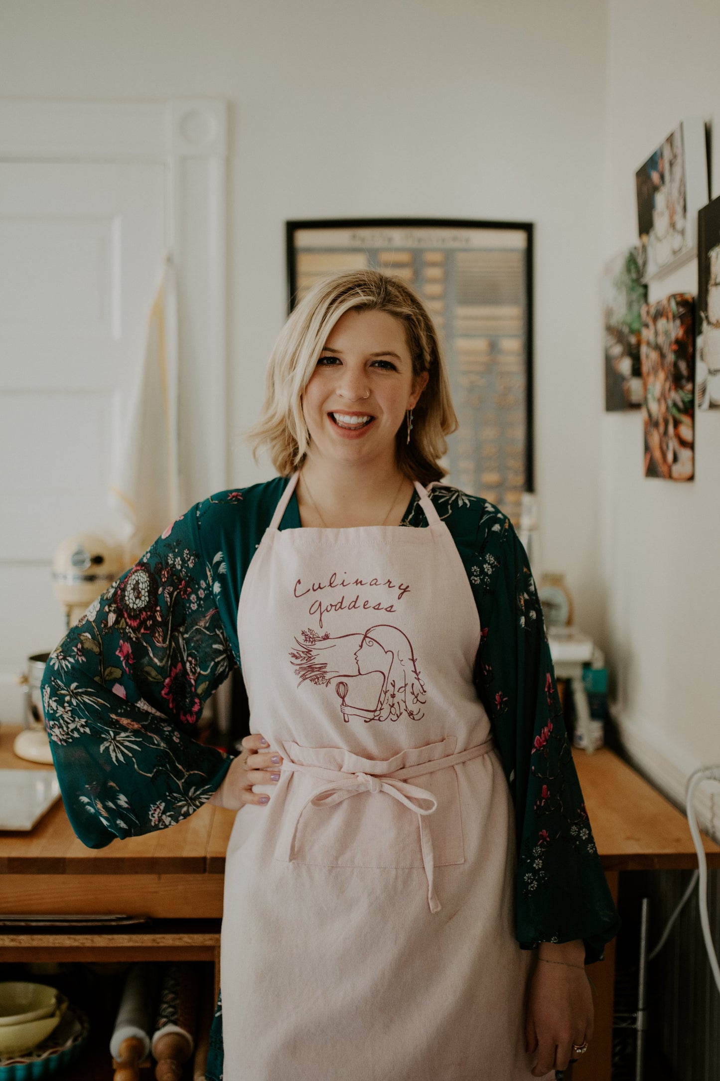 Woman in a pink apron that says "Culinary Goddess" stands in her kitchen