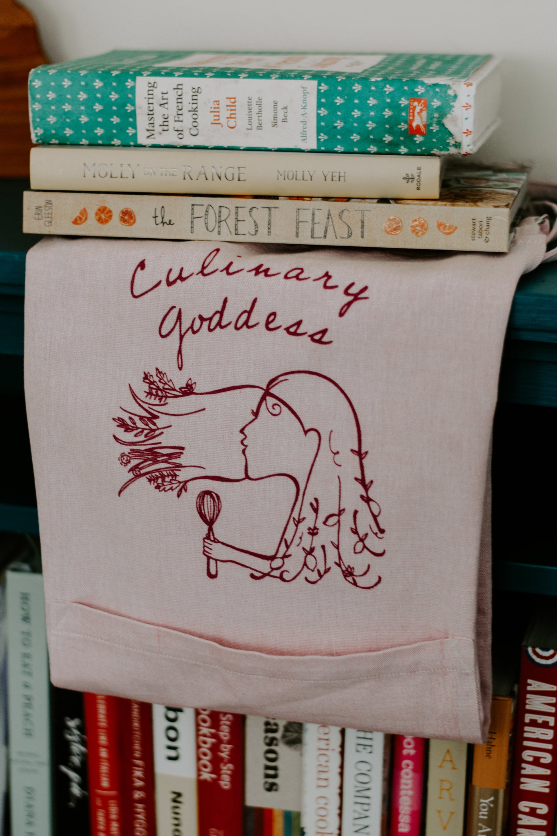 Pink Culinary Goddess apron folded and hung with cookbooks