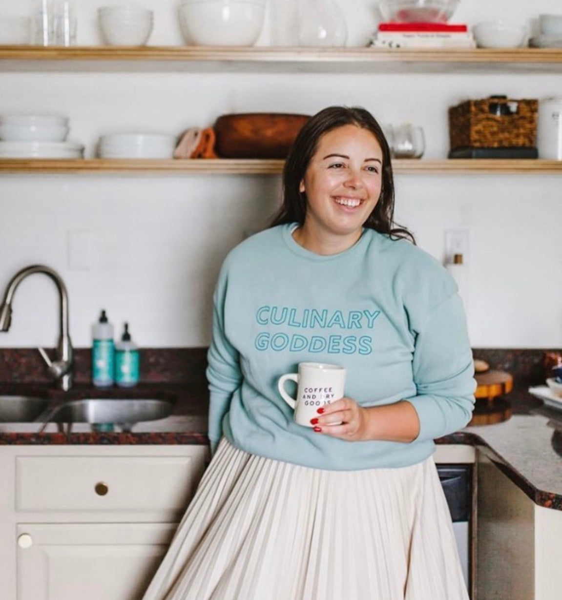 Woman holding coffee cup in her kitchen wearing dusty blue Culinary Goddess crewneck