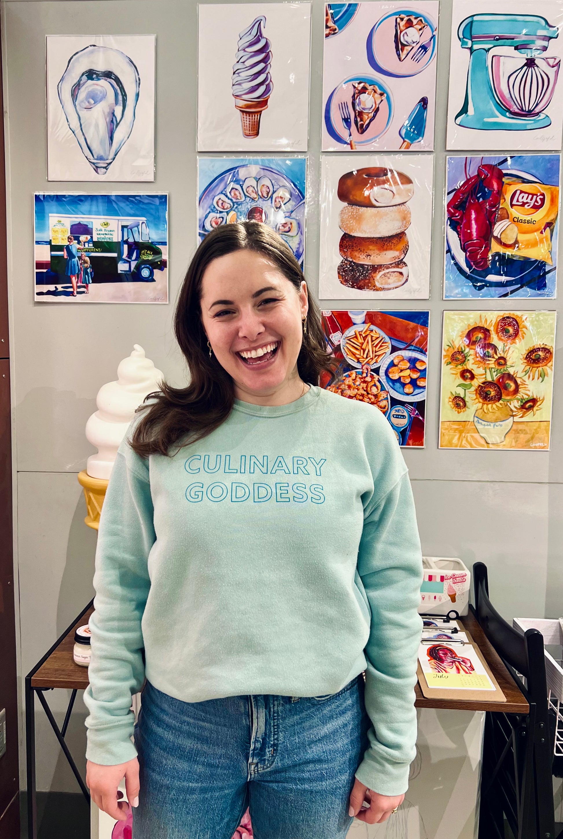 Woman in blue Culinary Goddess crewneck standing in front of food illustrations