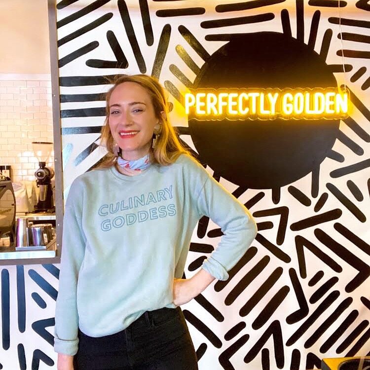 Woman wearing Culinary Goddess sweatshirt in front of a sign that says "Perfectly Golden"