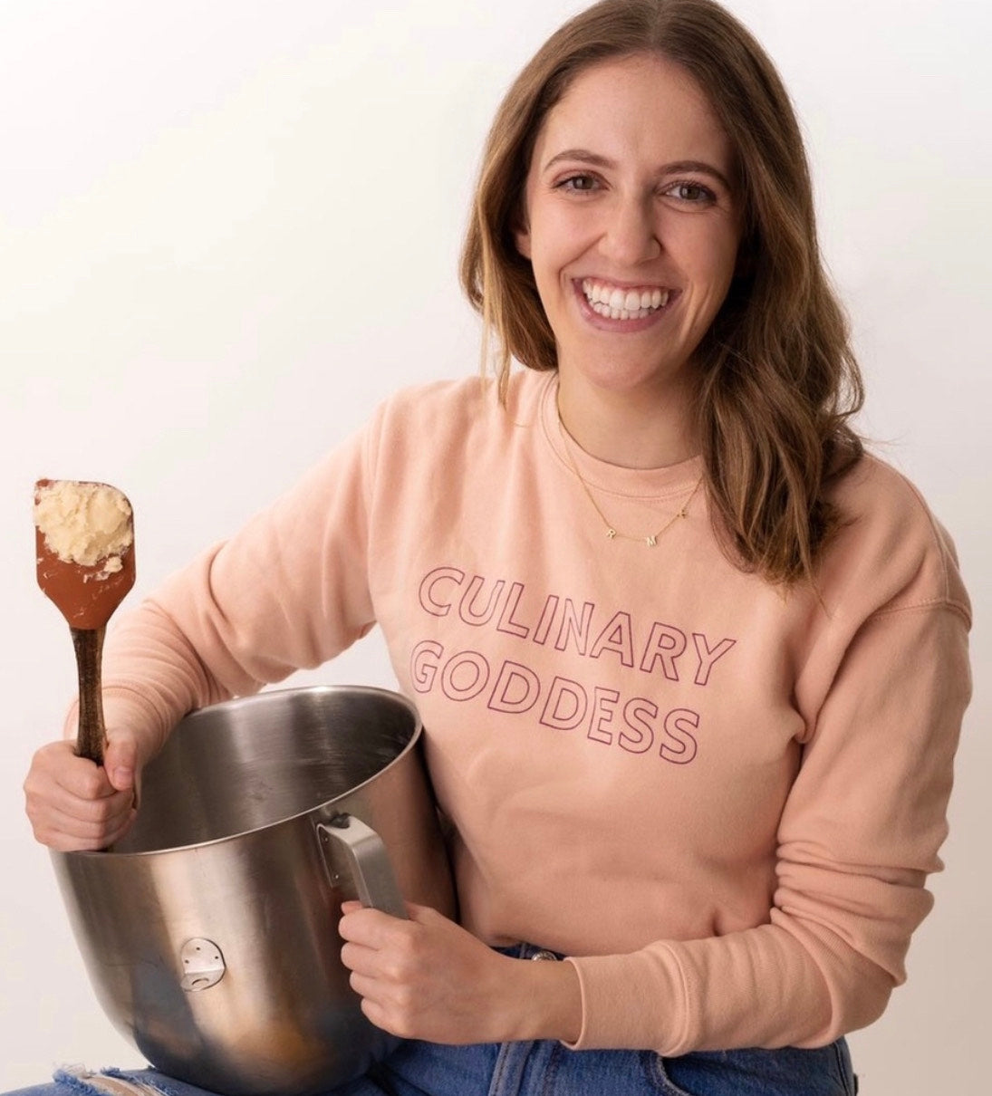 A woman in a pink Culinary Goddess sweatshirt smiling and holding a mixing bowl and spatula