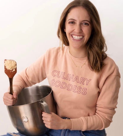 A woman in a pink Culinary Goddess sweatshirt smiling and holding a mixing bowl and spatula