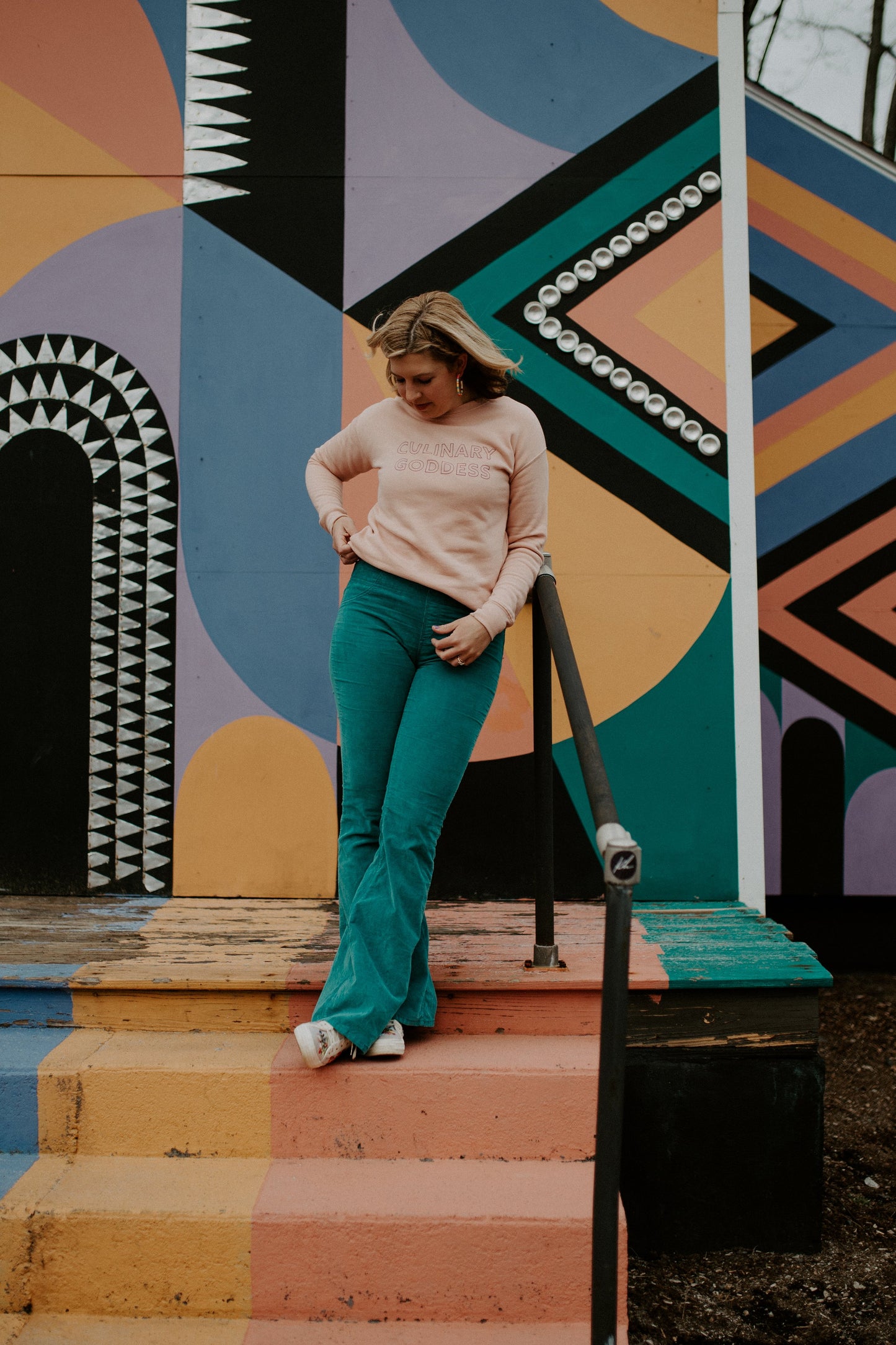 Woman in pink Culinary Goddess sweatshirt and teal pants in front of a colorful mural