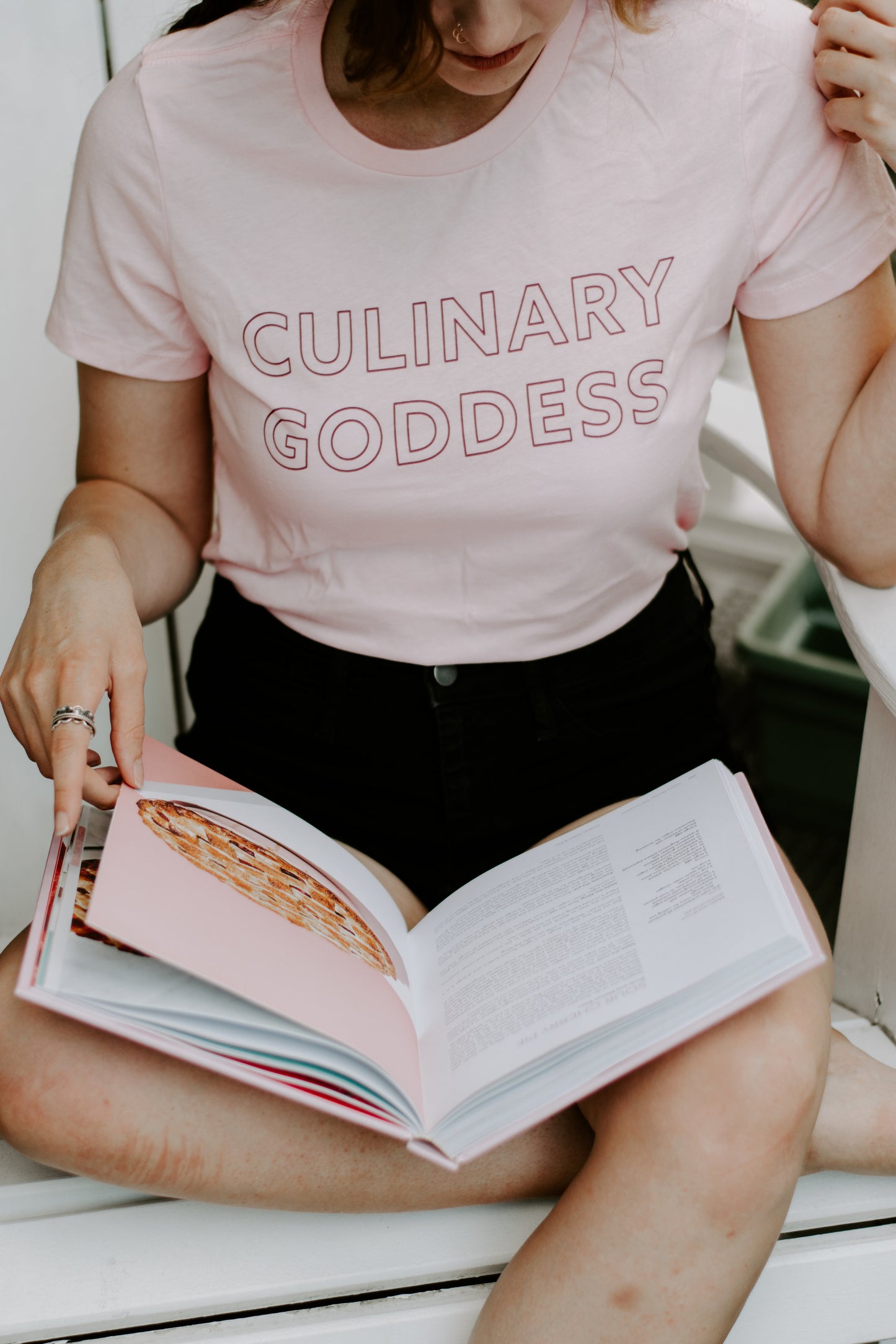 A woman wearing a pink Culinary Goddess tee with black shorts reading a cookbook