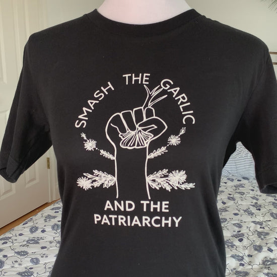 A black tee with the words "Smash the Garlic and the Patriarchy" and an illustration of a hand holding garlic hangs on a manikin