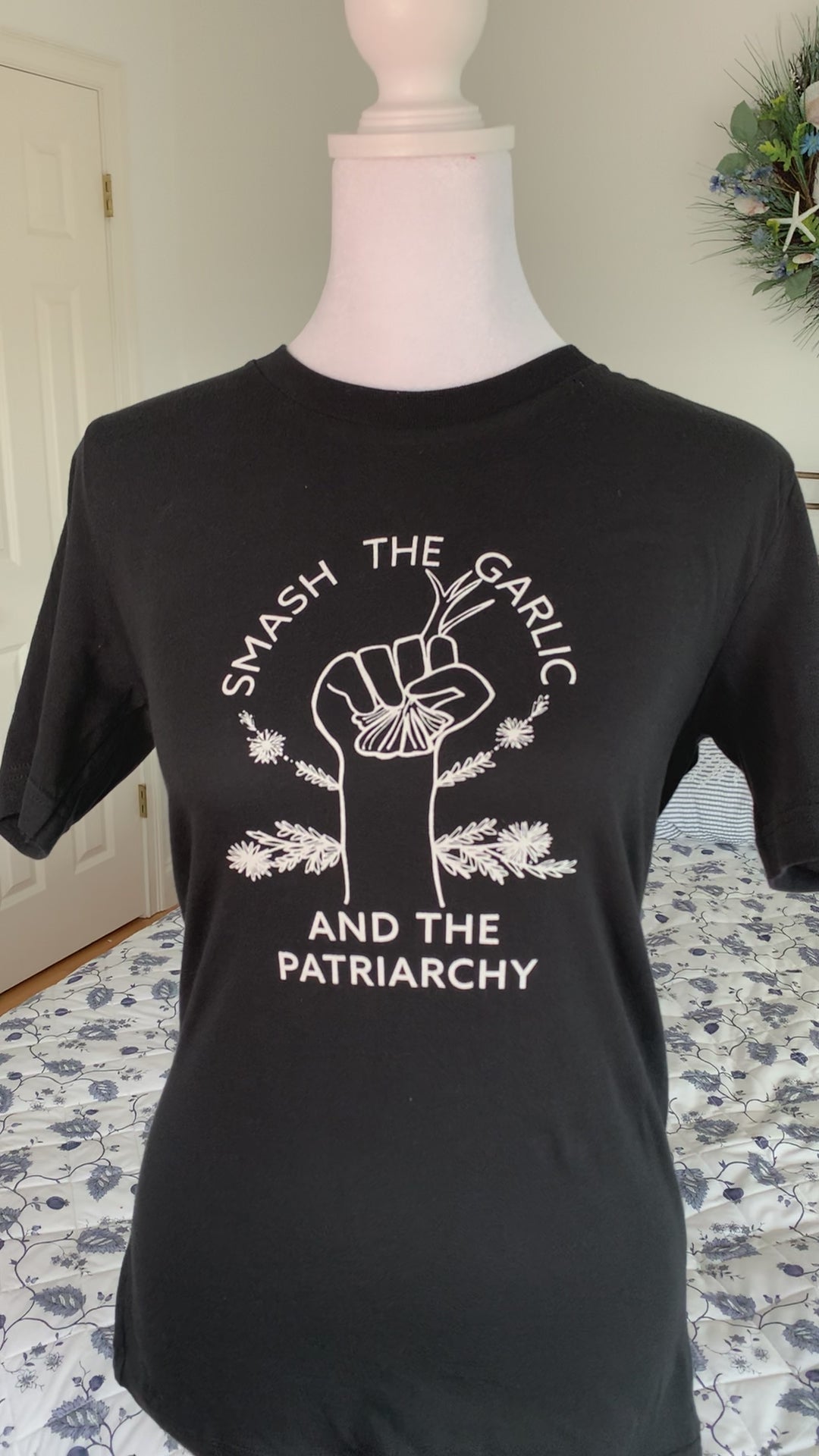A black tee with the words "Smash the Garlic and the Patriarchy" and an illustration of a hand holding garlic hangs on a manikin