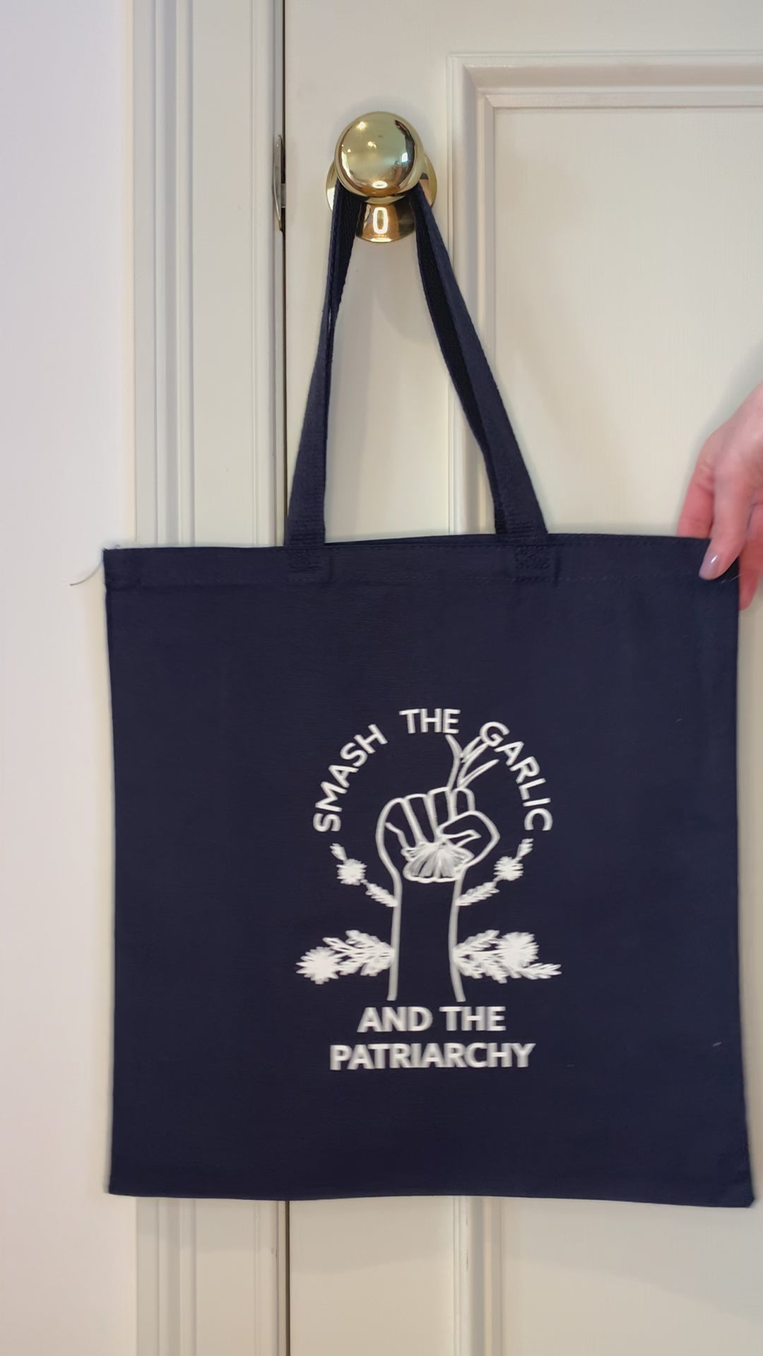 A navy blue tote that reads "Smash the Garlic and the Patriarchy" hangs on a doorknob