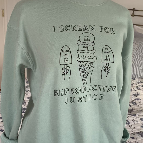 A dusty blue crewneck sweatshirt that reads "I scream for reproductive justice" hangs on a manikin
