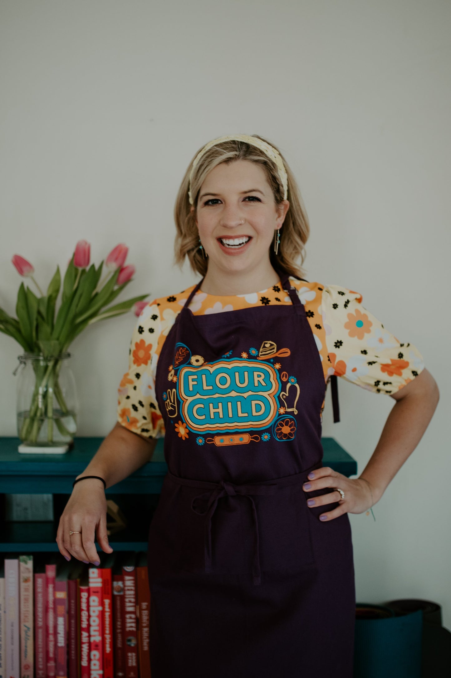 A woman wears a purple apron with the words "Flour Child" and colorful kitchen illustrations over a bright patterned top