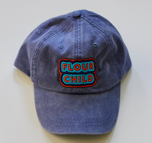 A blue baseball cap with the words Flour Child written in block letters with a blue, purple and orange outline
