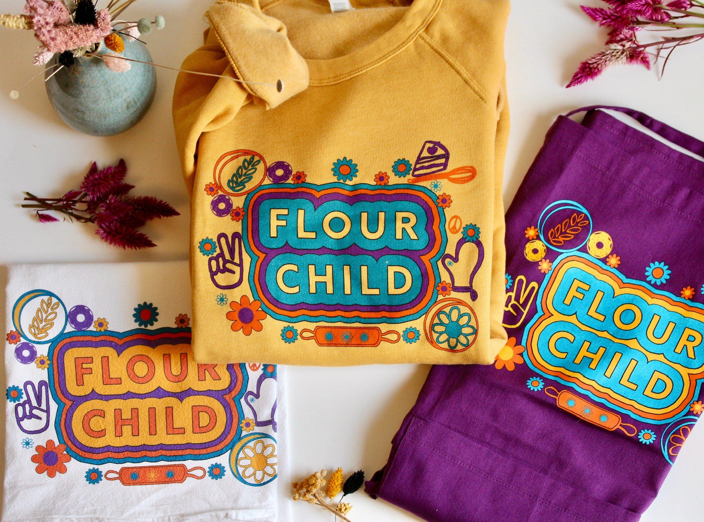 A white tea towel with the words Flour Child, a yellow crewneck with the words Flour Child and a purple Flour Child apron