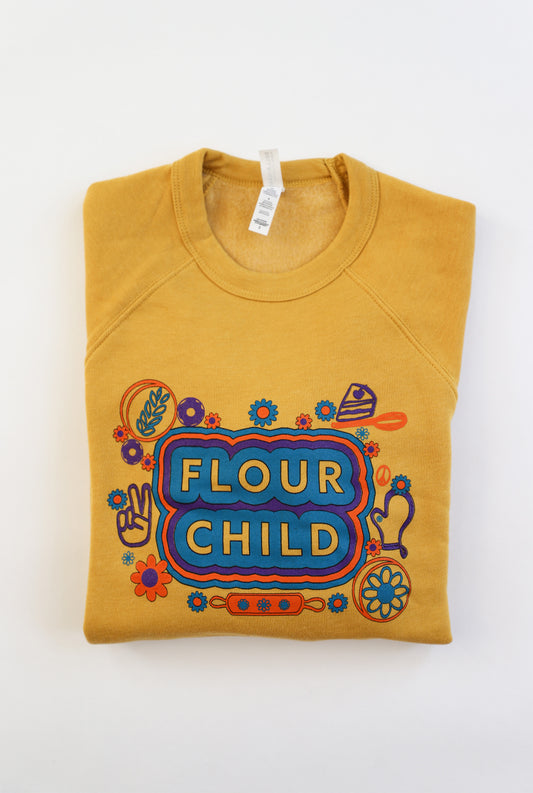A folded yellow crewneck that has the words "Flour Child" and sweet kitchen illustrations in blue, purple and orange.